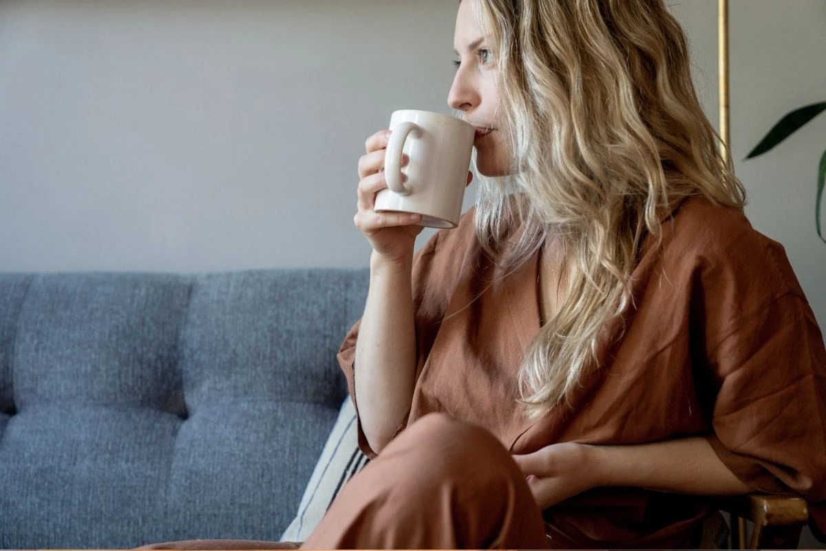 Kobieta z długimi blond włosami siedzi na szarej kanapie i pije napój z białego kubka, ubrana w brązowy, wygodny strój domowy, spogląda w bok, pochłonięta własnymi myślami.