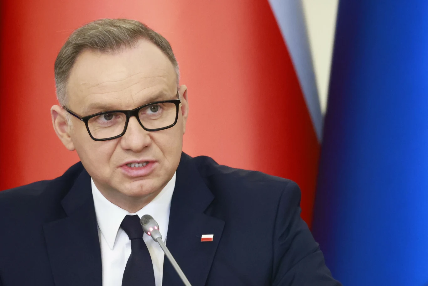 Andrzej Duda. Zdj. ilustracyjne Mężczyzna w garniturze i okularach mówi do mikrofonu na tle flag o kolorach czerwonym, białym i niebieskim, po lewej stronie widoczna przypinka w barwach narodowych.