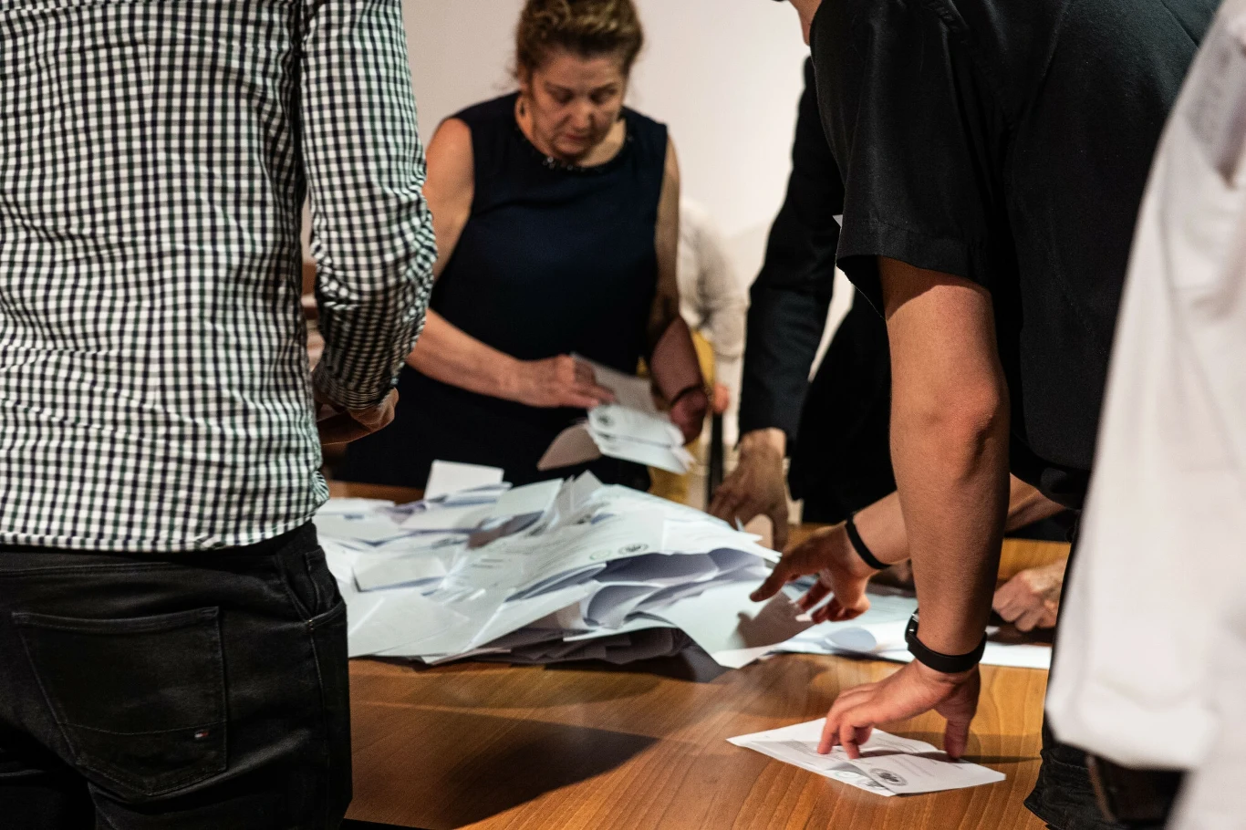 Liczenie głosów po drugiej turze wyborów prezydenckich Kilka osób stoi przy stole i przegląda stos papierów lub kopert, skupiając się na ich sortowaniu lub liczeniu.