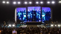 Tłum ludzi z telefonami uniesionymi do nagrywania występu na dużej scenie z jasnym oświetleniem, na scenie widoczny zespół muzyczny oraz ogromny ekran wyświetlający artystów i efekty wizualne.