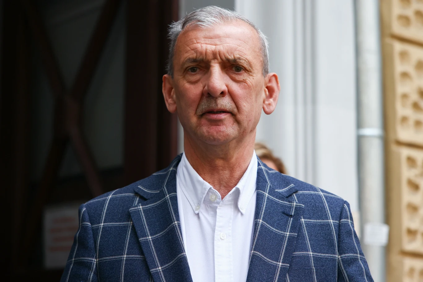 Sławomir Broniarz, prezes ZNP Starszy mężczyzna w eleganckiej marynarce w kratę i białej koszuli stoi przed wejściem do budynku z kamiennymi elementami fasady.