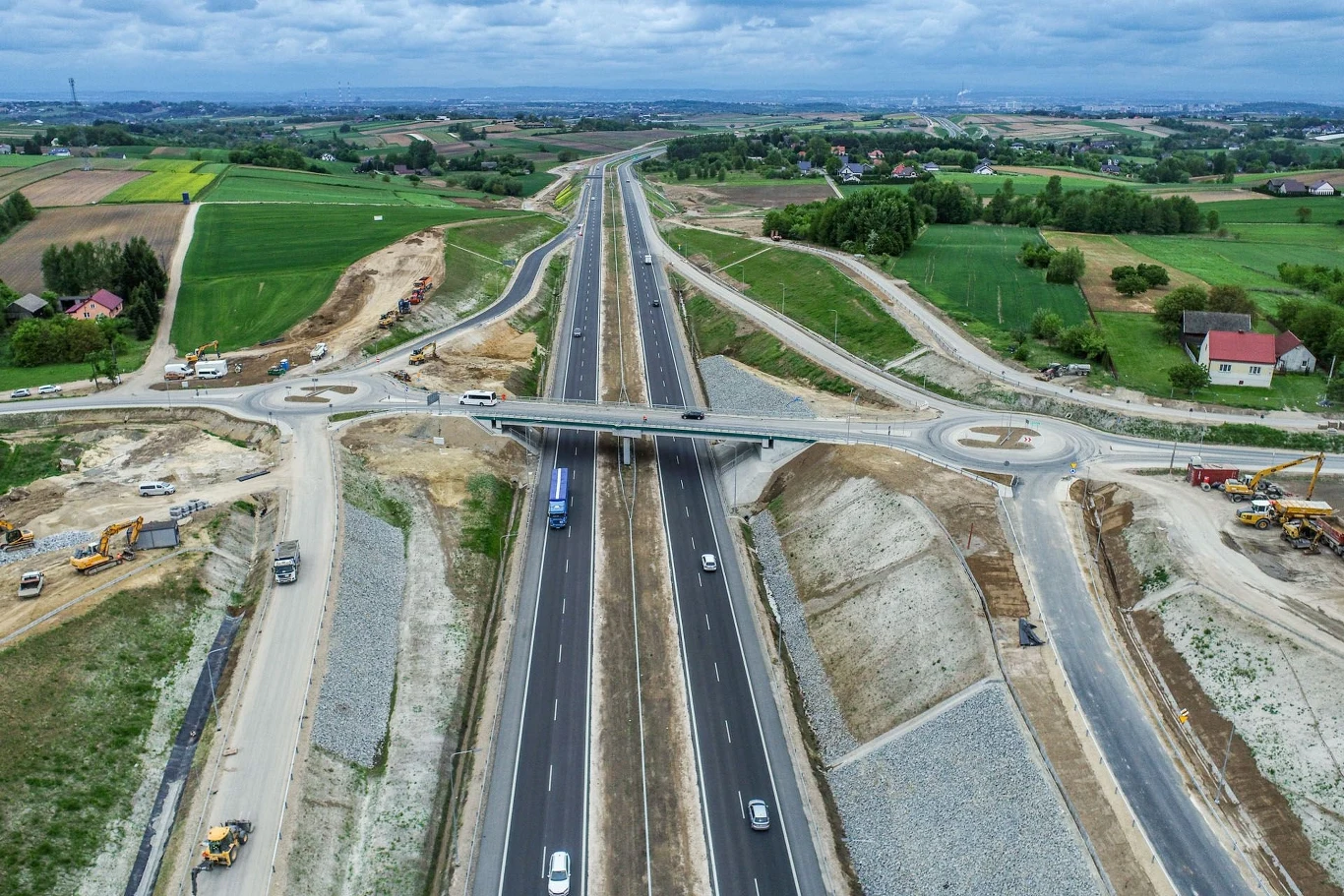 Rozbudowana dwupasmowa autostrada z wiaduktem nad jezdnią, otoczona zielonymi polami, widoczne są maszyny budowlane oraz kilka samochodów, w tle pola uprawne i zabudowania wiejskie.