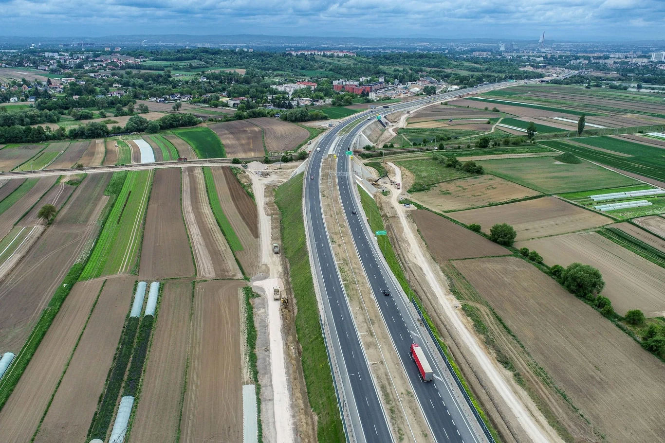 Nowo wybudowana droga ekspresowa biegnąca przez rozległe tereny rolnicze, widoczne pola uprawne po obu stronach trasy, pojedynczy samochód ciężarowy na jezdni, horyzont z zabudowaniami miejskimi oraz fragmentem lasu.
