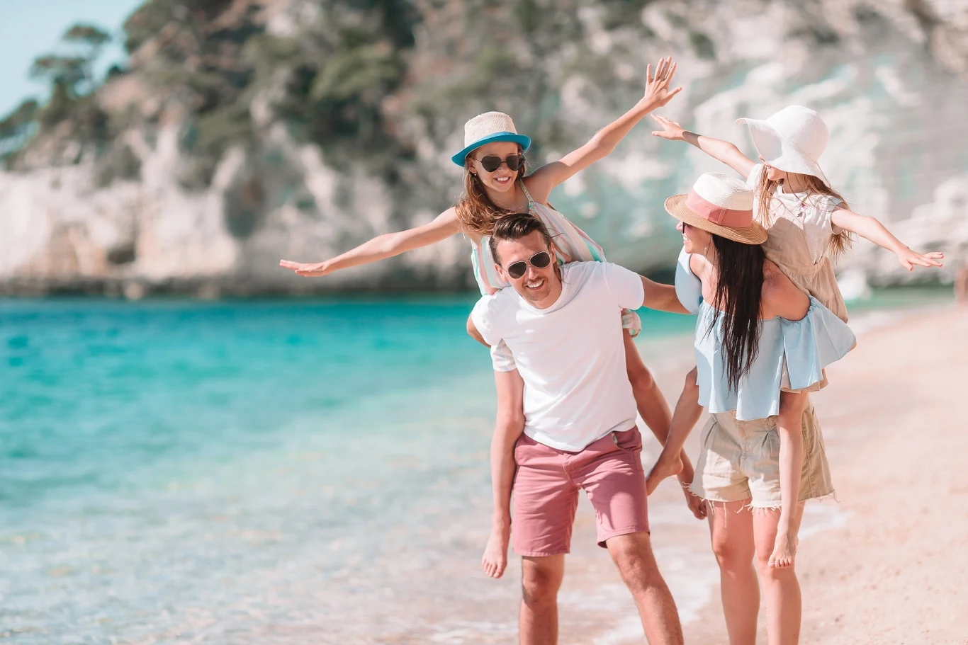 W ferworze przygotowań do wakacji łatwo zapomnieć o pewnej ważnej rzeczy Rodzina bawi się razem na piaszczystej plaży nad turkusową wodą, dwie dziewczynki mają słomkowe kapelusze, jedna siedzi na barana u dorosłego mężczyzny, wokół radosna atmosfera letniego wypoczynku.