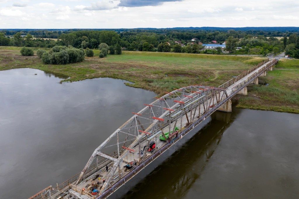 Cigacice , remontowany most nad Odra rok 2020 Stalowy most przebiegający nad szeroką rzeką, z widocznymi pracami remontowymi oraz maszynami na powierzchni konstrukcji, otoczony zielonymi łąkami i drzewami w tle.