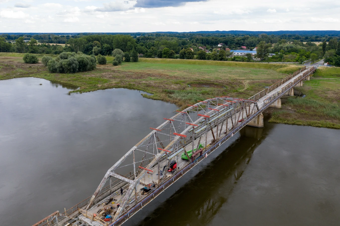 Cigacice , remontowany most nad Odra rok 2020 Stalowy most przebiegający nad szeroką rzeką, z widocznymi pracami remontowymi oraz maszynami na powierzchni konstrukcji, otoczony zielonymi łąkami i drzewami w tle.