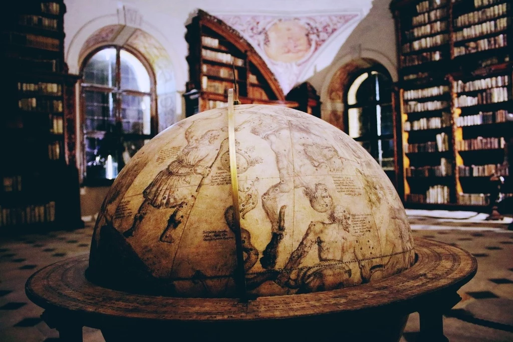 Żagańska biblioteka klasztorna Duży, antyczny globus astronomiczny z ilustracjami konstelacji, otoczony półkami pełnymi starych książek w stylowym, zabytkowym wnętrzu biblioteki.