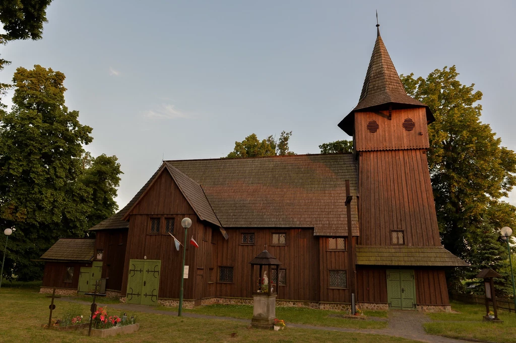 Chlastawa. Szlak Kościołów Drewnianych Regionu Kozla Drewniany kościół z wysoką wieżą, otoczony zielonymi drzewami i niewielkim ogrodem z kwiatami, widoczna flaga Polski zawieszona na elewacji budynku.