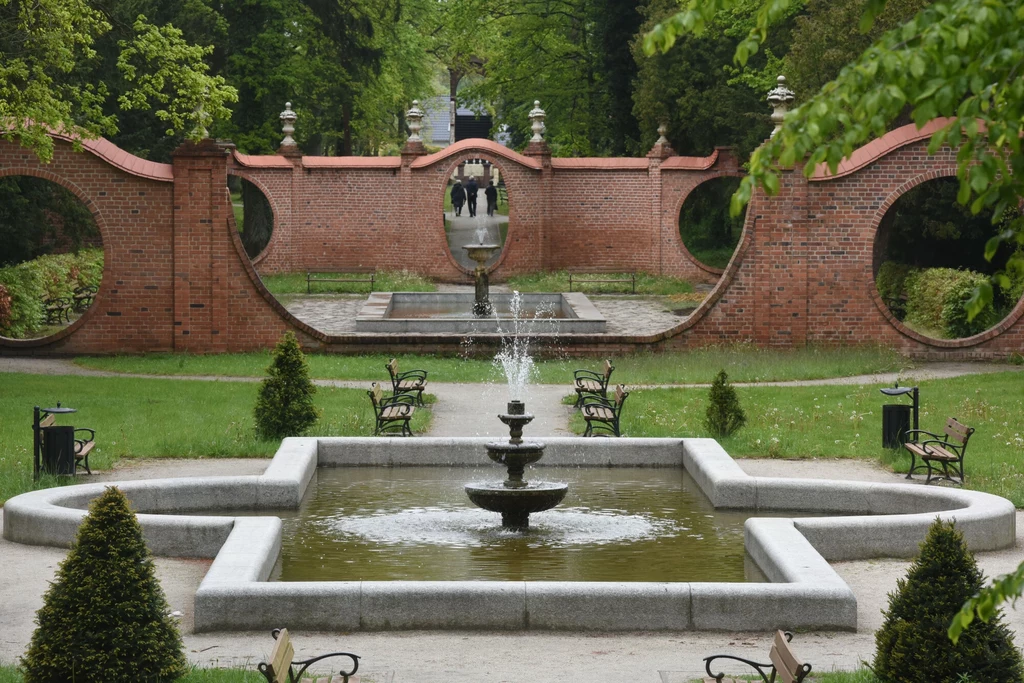 Park Dworski. Iłowa Żagańska Centralna fontanna otoczona zielonymi trawnikami i krzewami, naprzeciw efektownego, czerwonego muru z okrągłymi prześwitami, w tle widoczne ławki i alejki parkowe oraz spacerujące osoby.