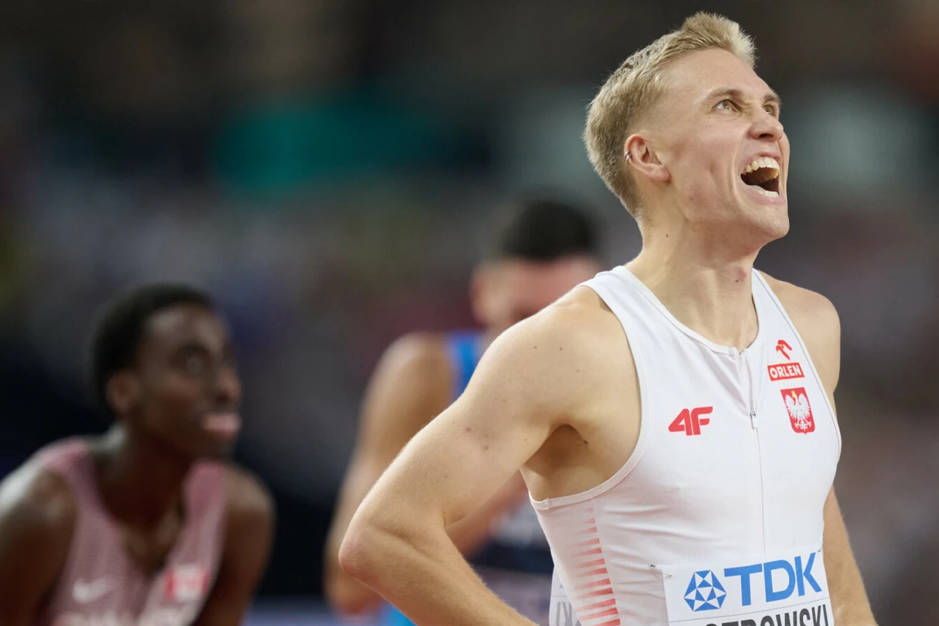 Lekkoatleta w białym stroju reprezentacji Polski podczas zawodów sportowych, z wyrazem radości na twarzy, w tle inni sportowcy na bieżni.