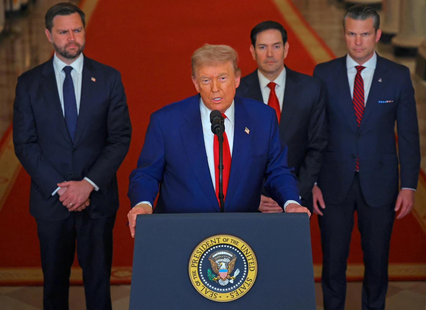 Donald Trump podczas przemówienia po atakach USA na obiekty nuklearne Iranu Mężczyzna w niebieskim garniturze stoi przy mównicy z pieczęcią prezydenta Stanów Zjednoczonych, po jego bokach stoją trzej inni mężczyźni w garniturach, w tle czerwony dywan i kolumny reprezentacyjne, atmosfera oficjalna.