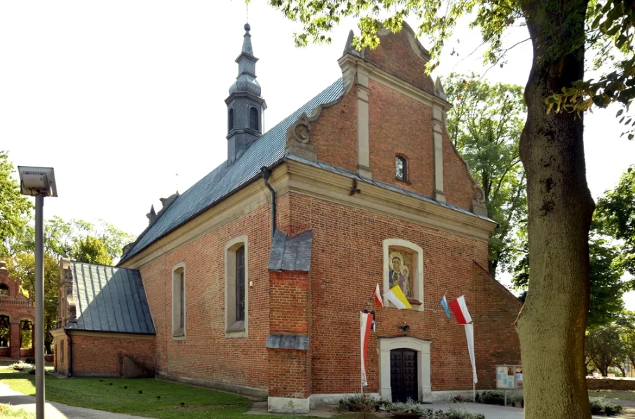 Ciało 31-latka odnaleziono na terenie parafii w Drobinie w 2024 roku Ceglany kościół z wieżą i frontonem, ozdobiony flagami Polski oraz obrazem nad wejściem, otoczony zielonymi drzewami i niskim ogrodzeniem, w słoneczny dzień.