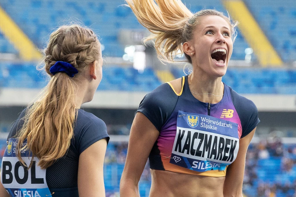 Natalia Bukowiecka i Femke Bol (tyłem) Dwie lekkoatletki na stadionie, jedna z nich z wyraźną radością na twarzy i tabliczką z nazwiskiem Kaczmarek na piersi, druga zwrócona do niej bokiem, w tle trybuny z publicznością.