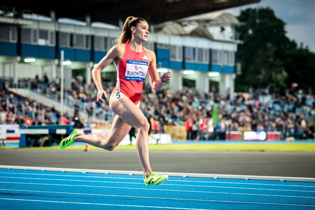 Lekkoatletka w czerwonym stroju sportowym biegnie po niebieskim torze podczas zawodów na stadionie, w tle widoczna publiczność na trybunach.