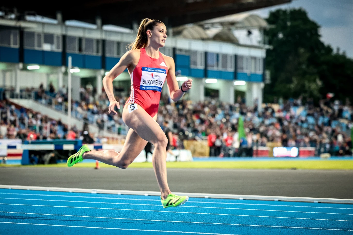Lekkoatletka w czerwonym stroju sportowym biegnie po niebieskim torze podczas zawodów na stadionie, w tle widoczna publiczność na trybunach.