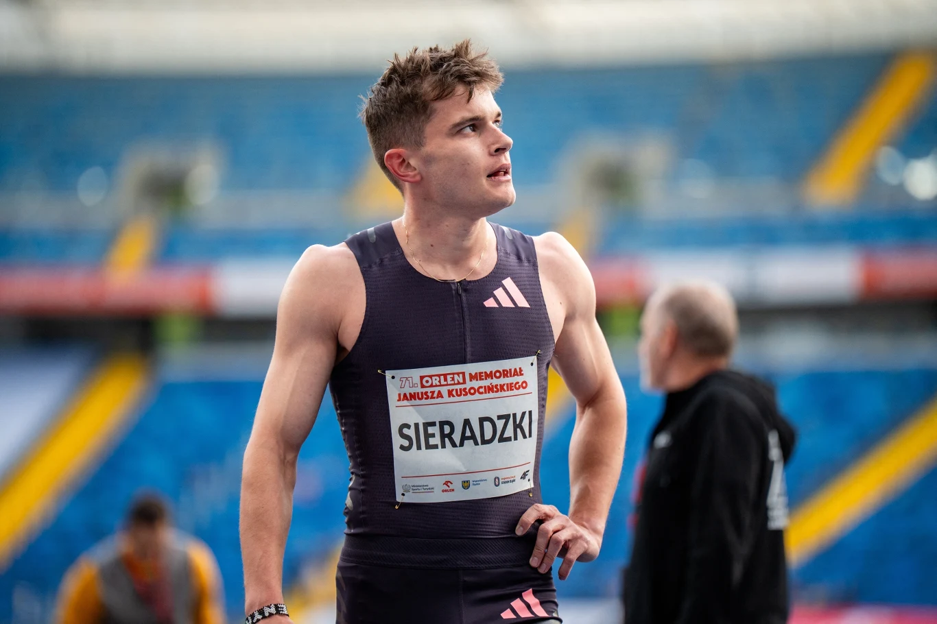 Sportowiec w sportowym stroju z numerem startowym Sieradzki stoi na stadionie lekkoatletycznym, w tle rozmyte sylwetki osób oraz niebieskie trybuny.