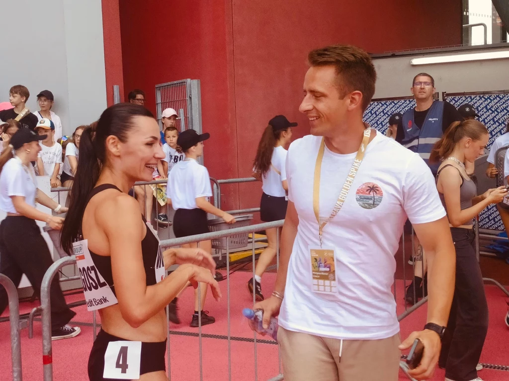 Sportsmenka w stroju startowym rozmawia z mężczyzną posiadającym akredytację, oboje uśmiechnięci, w tle widoczni wolontariusze i osoby obsługujące wydarzenie sportowe.
