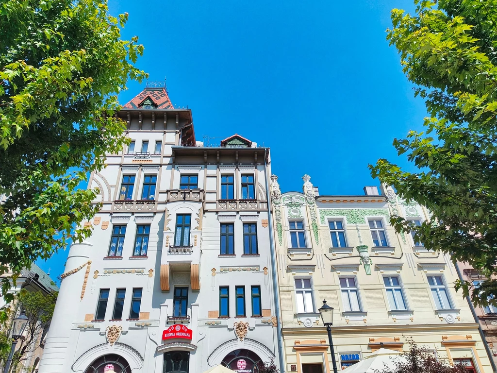 Odrestaurowane, zabytkowe kamienice z dekoracyjnymi fasadami, między nimi widoczne zielone korony drzew, na tle intensywnie niebieskiego nieba.