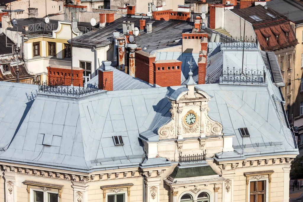 Zabytkowa kamienica z bogato zdobioną fasadą i wieżyczkami na dachu, ozdobiona zegarem, w tle widać dachy innych pobliskich budynków, charakterystyczne czerwone kominy i klimatyczne detale architektoniczne.
