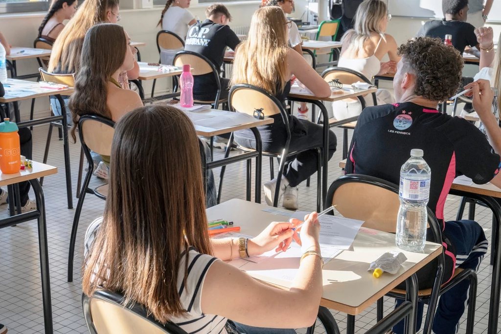 Sztuczna inteligencja coraz silniej wpływa na szkolnictwo wyższe Grupa młodych ludzi siedzi w sali lekcyjnej przy osobnych ławkach, skupiona na pisaniu testu lub egzaminu, na ławkach widoczne są kartki, długopisy oraz butelki z wodą.