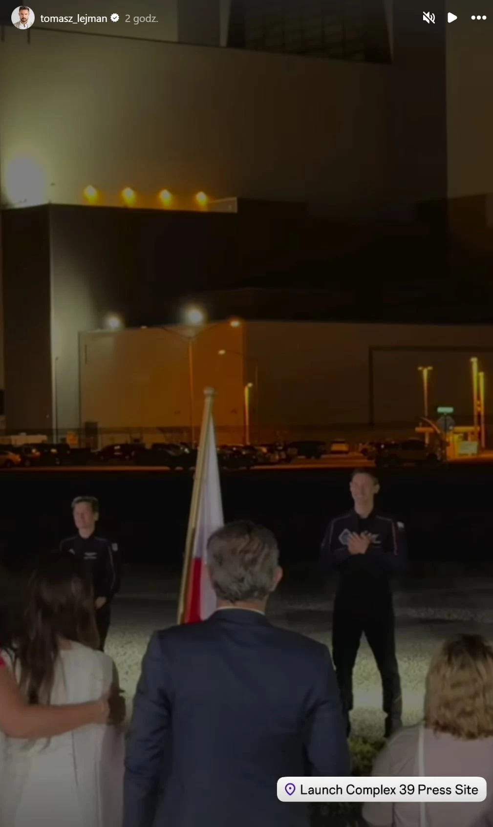 Pożegnanie Sławosza z rodziną Ceremonia z udziałem kilku osób, grupa stoi przed polską flagą na tle nocnego oświetlenia i nowoczesnego budynku, w tle strefa przemysłowa oraz wejście do kompleksu.