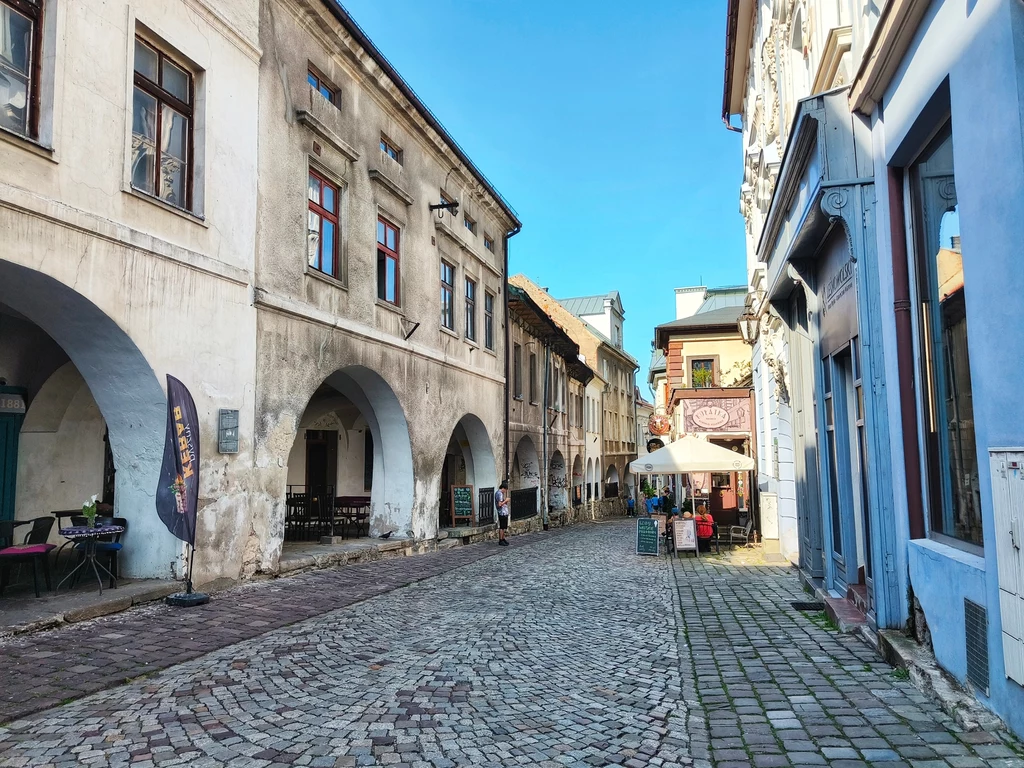 Wąska brukowana uliczka z zabytkową zabudową po obu stronach, liczne łuki i arkady, stoliki pod parasolami ustawione przy jednej z kamienic, bezludna, jasna scena w słoneczny dzień.
