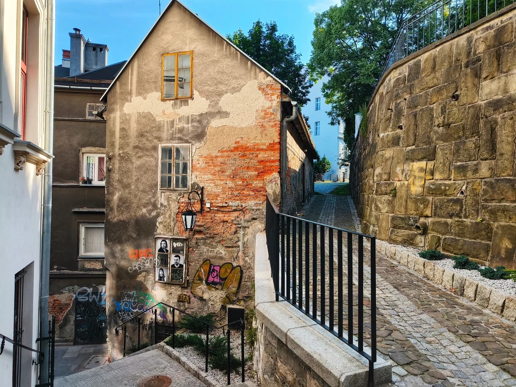 Stara kamienica z widocznymi zniszczeniami elewacji, częściowo odsłoniętą cegłą i graffiti na murze, obok brukowana uliczka ze wzniesieniem oraz kamienny mur oporowy, w tle roślinność i drzewa.