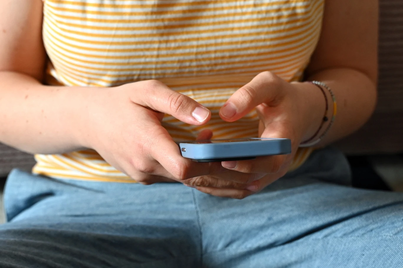 Ręce osoby trzymającej smartfon, ubranej w żółtą bluzkę w paski i niebieskie spodnie, na nadgarstku widoczna bransoletka.