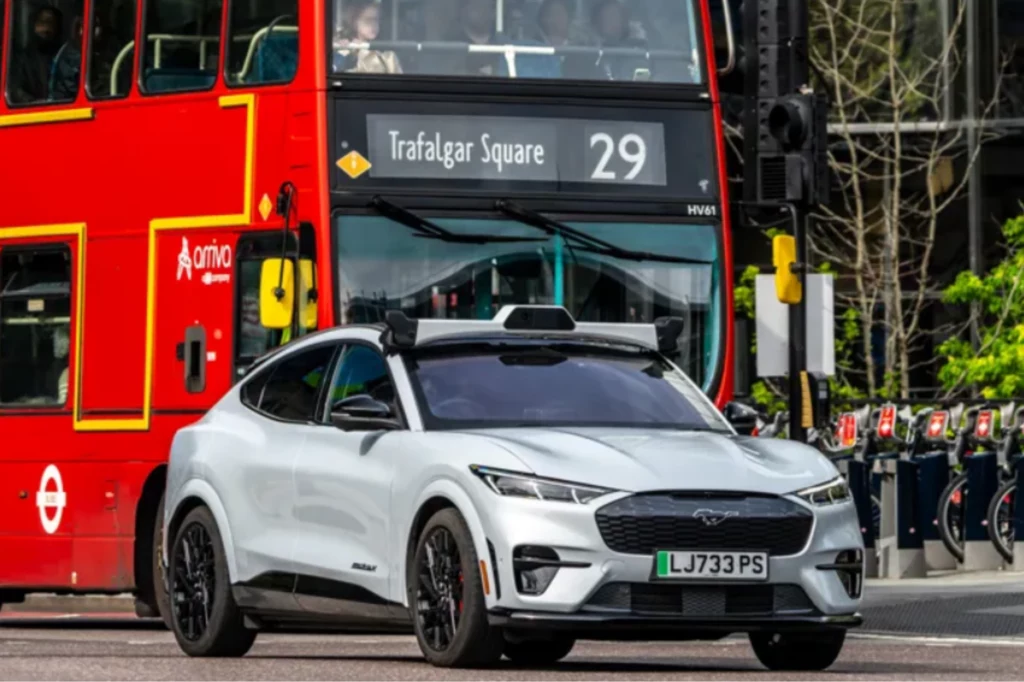 Uber podpisał współprace z firmą Wayve, która zajmuje się sztuczną inteligencją. Białe auto elektryczne marki Ford Mustang Mach-E z zamontowanym urządzeniem na dachu stoi na ulicy, w tle czerwony dwupiętrowy autobus londyński linii 29 z kierunkiem Trafalgar Square.