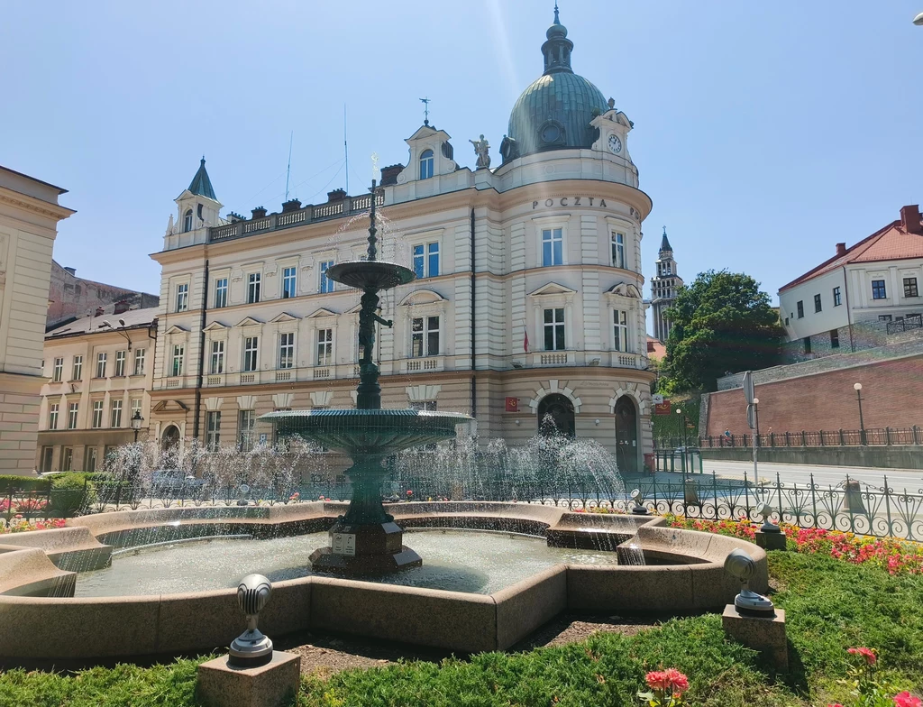Zabytkowy budynek o klasycystycznej architekturze z charakterystyczną kopułą, przed którym znajduje się okazała fontanna otoczona kwiatami i zielenią, w tle widoczne są inne budynki miejskie.