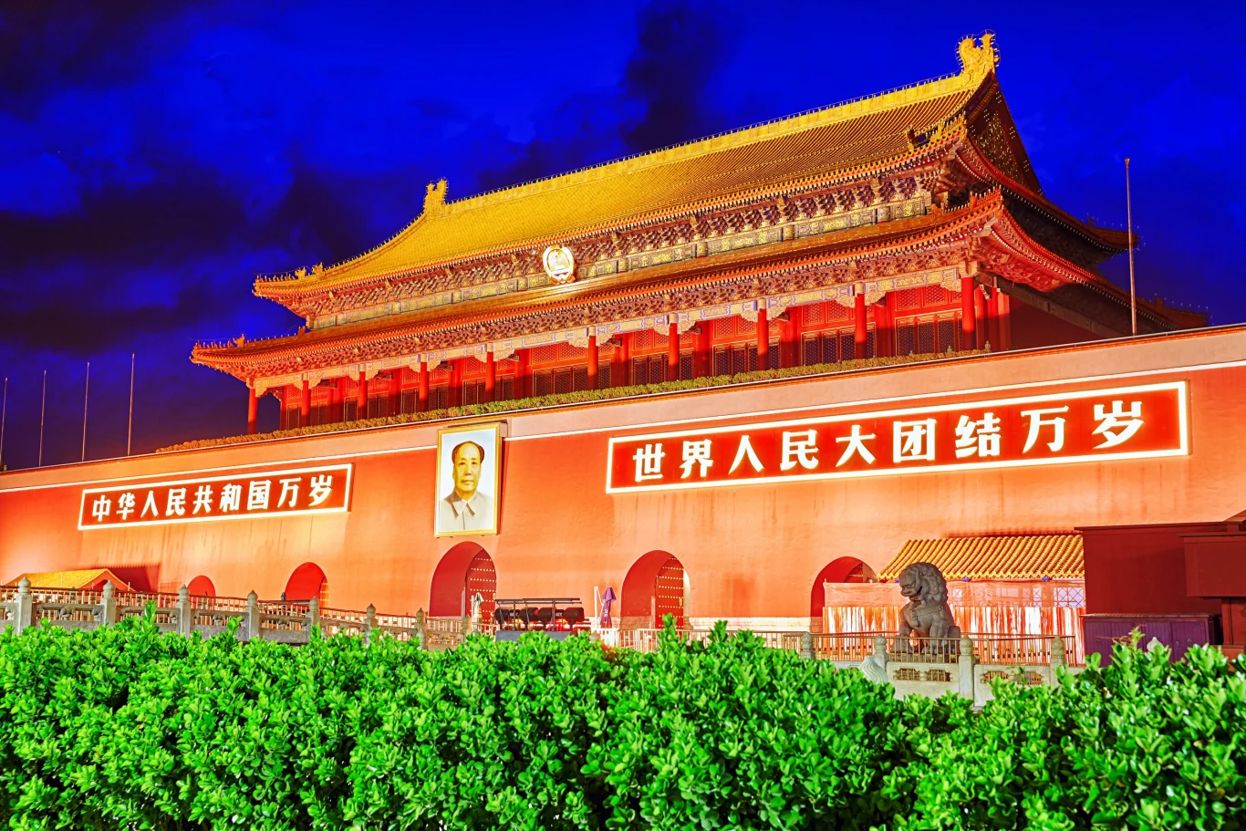 Oświetlona brama Tiananmen w Pekinie nocą, z portretem Mao Zedonga pośrodku i czerwonymi banerami z chińskimi napisami na elewacji, otoczona zielonymi krzewami oraz rzeźbami lwów przed wejściem.