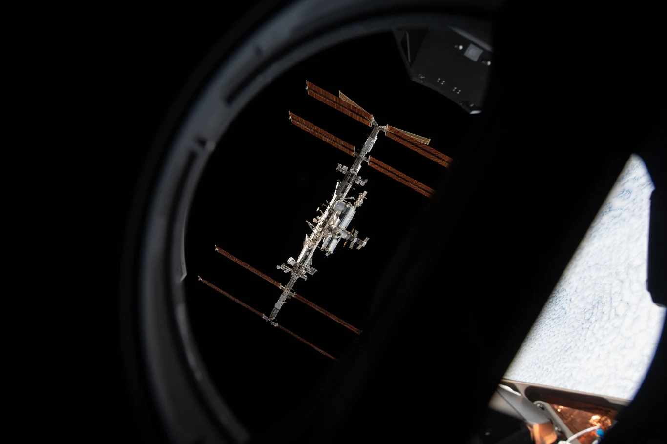 Międzynarodowa stacja kosmiczna ISS - widok z kapsuły Dragon Międzynarodowa Stacja Kosmiczna widziana przez okno statku kosmicznego na tle czerni kosmosu, jasno oświetlona konstrukcja z charakterystycznymi panelami słonecznymi.
