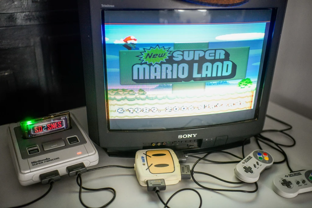 Stara konsola Nintendo Stary telewizor Sony z włączoną grą New Super Mario Land, konsola Super Nintendo Entertainment System (Super NES) z przystawką i podłączonymi dwoma kontrolerami, utrzymane w klimacie retro.