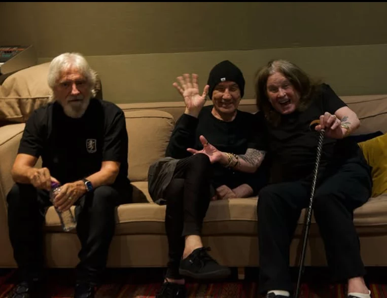 Od lewej Geezer Butler, Bill Ward, Ozzy Osbourne Trzech starszych mężczyzn siedzi na beżowej sofie, wszyscy uśmiechają się i sprawiają wrażenie zrelaksowanych. Jeden z nich macha do kamery, drugi opiera się o laskę, trzeci trzyma butelkę wody. Ubrani są na czarno, tło stanowi prosta ściana i kilka po...