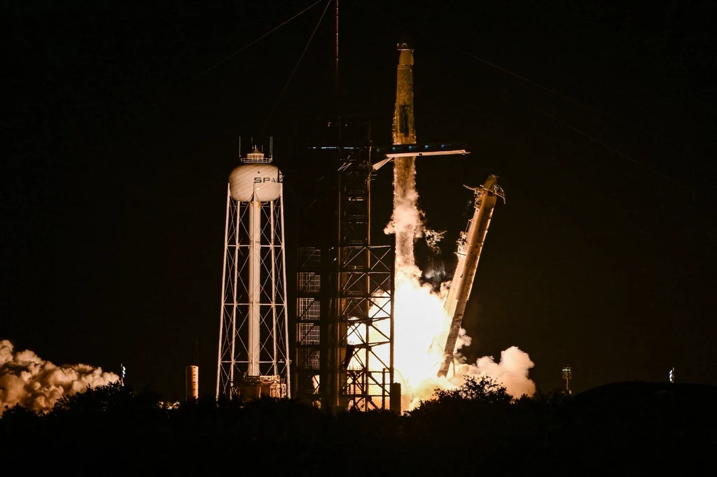 Rakieta startuje z platformy startowej nocą, widoczny intensywny płomień i dym, obok znajduje się wieża serwisowa ze zbiornikiem wody i logo SpaceX.