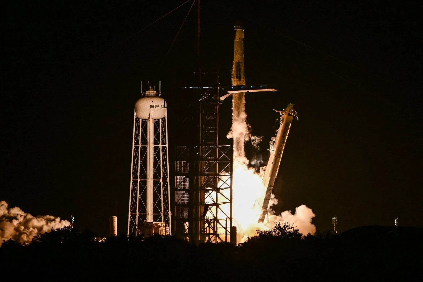 Rakieta startuje z platformy startowej nocą, widoczny intensywny płomień i dym, obok znajduje się wieża serwisowa ze zbiornikiem wody i logo SpaceX.