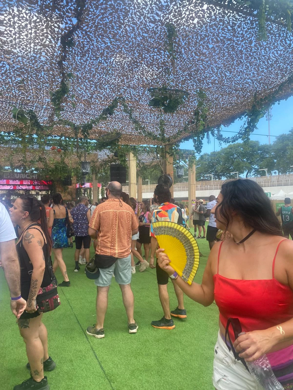 Barcelona Ludzie bawiący się na plenerowej imprezie pod dekoracyjnym dachem z siatki, jedna kobieta w czerwonym topie trzyma wachlarz i patrzy na grupę zgromadzoną przy scenie; wokół panuje słoneczna pogoda.