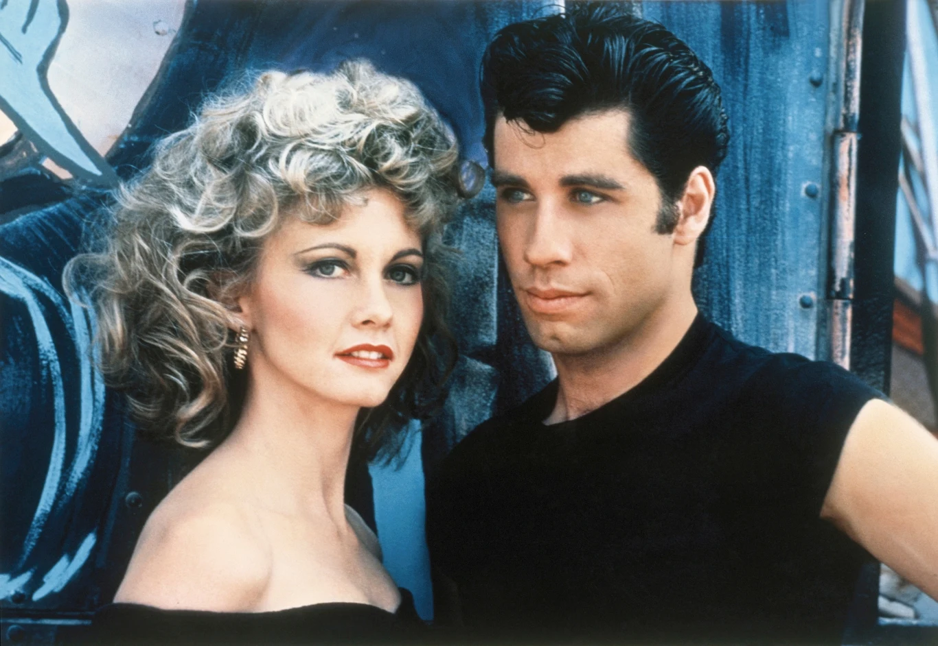 Olivia Newton-John i John Travolta na planie filmu "Grease" w 1978 r. Kobieta z jasnymi, kręconymi włosami i wyrazistym makijażem stoi obok mężczyzny z ciemnymi, uczesanymi do tyłu włosami ubranego w czarną koszulkę, oboje pozują zwróceni w stronę kamery na tle muralu.