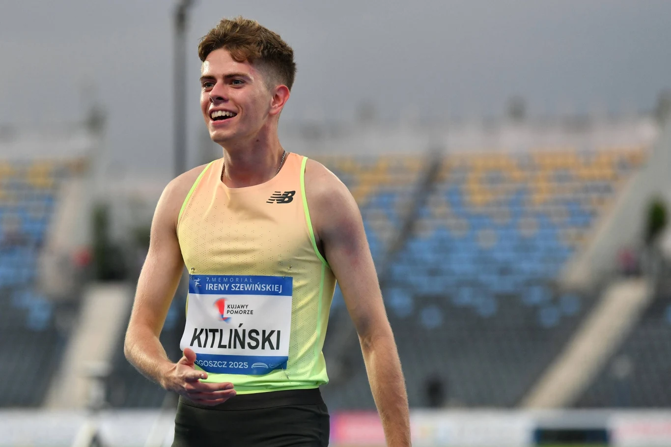 Bartosz Kitliński Młody lekkoatleta w jasnej koszulce i z numerem startowym, uśmiecha się po zakończonym biegu, w tle trybuny stadionu z miejscami dla widzów.