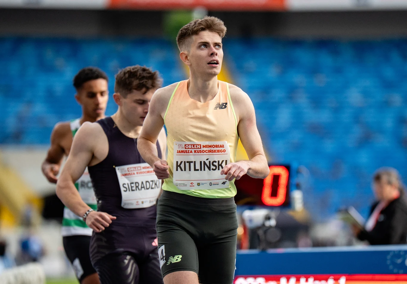 Bartosz Kitliński Zawodnik biegnący na stadionie lekkoatletycznym w trakcie zawodów, na koszulce nazwisko Kitliński, w tle inni biegacze oraz widownia na niebieskich trybunach.