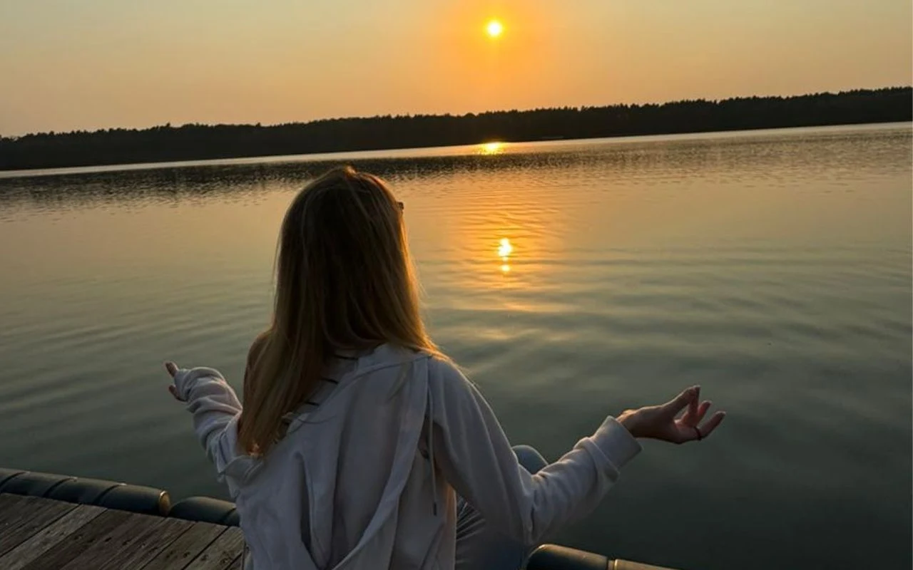 Kobieta siedząca na pomoście zwrócona w stronę spokojnego jeziora podczas zachodu słońca, dłonie ułożone w geście medytacji, w tle linią horyzontu przebiega zalesiony brzeg.