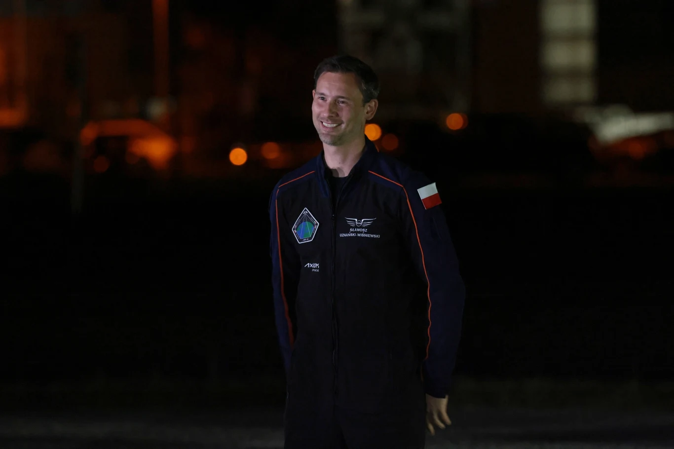 Politycy gratulują Sławoszowi Uznańskiemu-Wiśniewskiemu po starcie w kosmos. Na zdjęciu: Polski astronauta w bazie NASA na Florydzie na dobę przed rozpoczęciem misji kosmicznej Mężczyzna w kombinezonie astronauty z naszywką polskiej flagi stoi na ciemnym tle oświetlonym punktowymi źródłami światła, uśmiecha się.