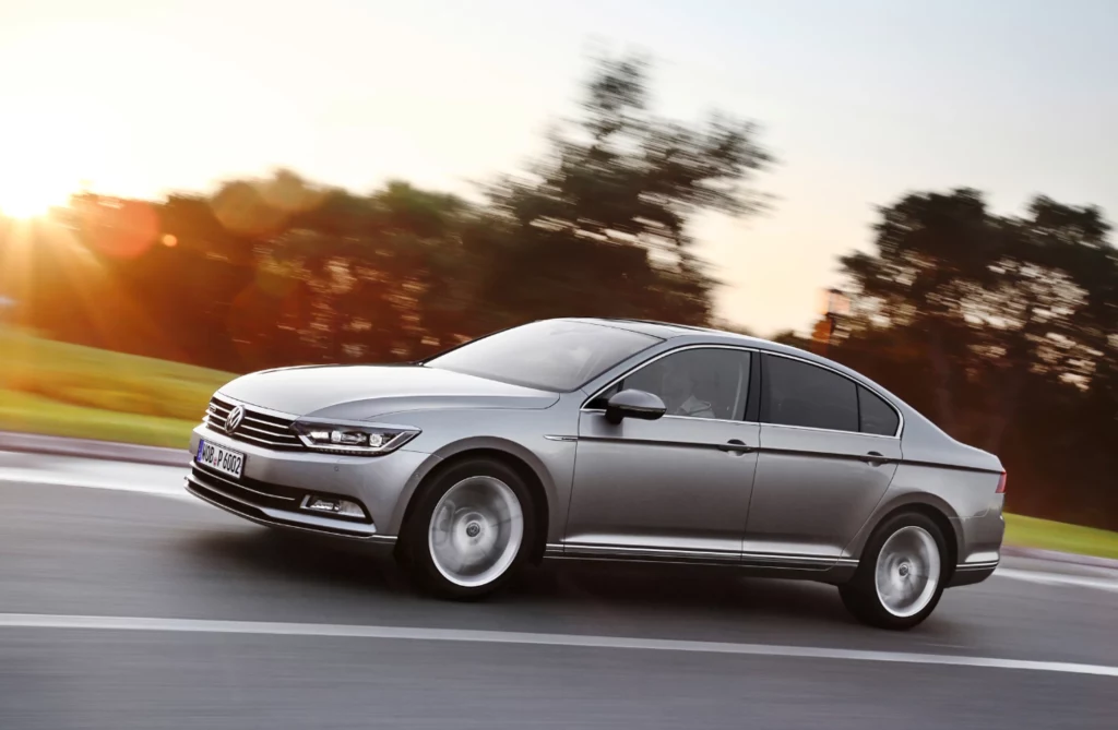 Sławosz Uznański-Wiśniewski na co dzień jeździ Volkswagenem Passatem B8. Srebrny samochód osobowy marki Volkswagen Passat porusza się po szerokiej, asfaltowej drodze, w tle rozmyte drzewa oraz jasne światło zachodzącego słońca.
