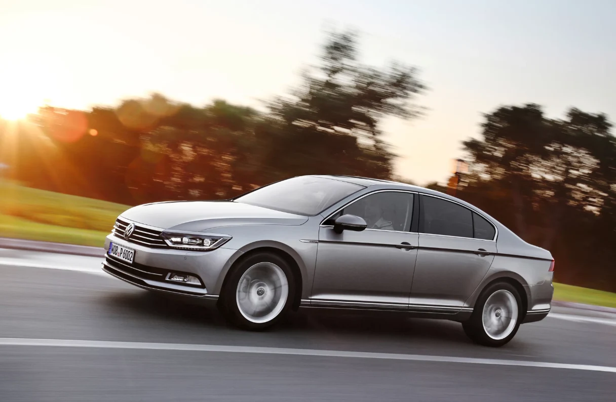 Sławosz Uznański-Wiśniewski na co dzień jeździ Volkswagenem Passatem B8. Srebrny samochód osobowy marki Volkswagen Passat porusza się po szerokiej, asfaltowej drodze, w tle rozmyte drzewa oraz jasne światło zachodzącego słońca.
