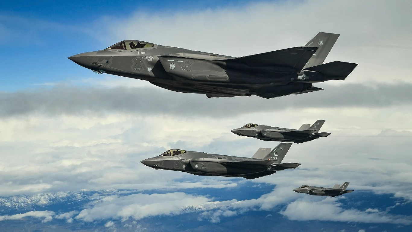 Cztery nowoczesne myśliwce wojskowe F-35 lecą w formacji nad chmurami na tle nieba, w oddali widoczne są ośnieżone szczyty gór.