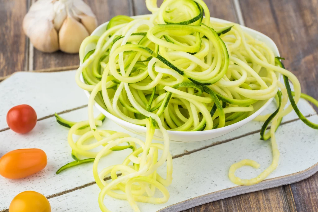 Cukinię łatwo zamienisz w lekki i zdrowy makaron Biała miska wypełniona spiralnie pokrojonymi surowymi warzywami przypominającymi makaron, obok znajdują się kolorowe małe pomidorki oraz główka czosnku na białej desce, wszystko na rustykalnym drewnianym tle.