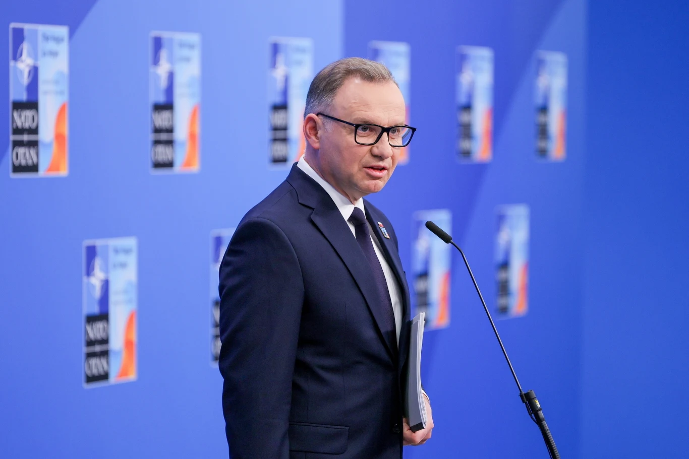 Szczyt NATO. Andrzej Duda zabrał głos Mężczyzna w garniturze i okularach stoi przy mównicy na tle niebieskiego panelu z logo NATO, trzymając w ręku dokumenty.