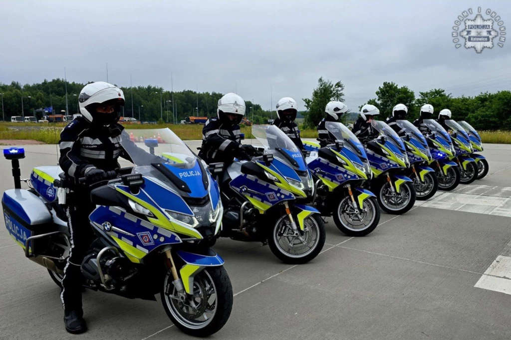 Motocyklowa Asysta Honorowa systematycznie trenuje i podnosi swoje umiejętności jazdy. Tym razem ćwiczenia odbywały się na autostradzie A4 oraz placu należącym do Komisariatu Autostradowej Policji w Gliwicach. Sześciu policjantów w pełnym umundurowaniu siedzi na nowych, niebieskich motocyklach policyjnych na parkingu, maszyny ustawione są w jednej linii, w tle widoczne drzewa i pole.