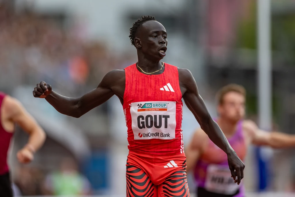 Gout Gout Sportowiec w czerwonym stroju startowym przekracza linię mety podczas zawodów lekkoatletycznych, wokół widoczni są inni biegacze i rozmyte tło stadionu.