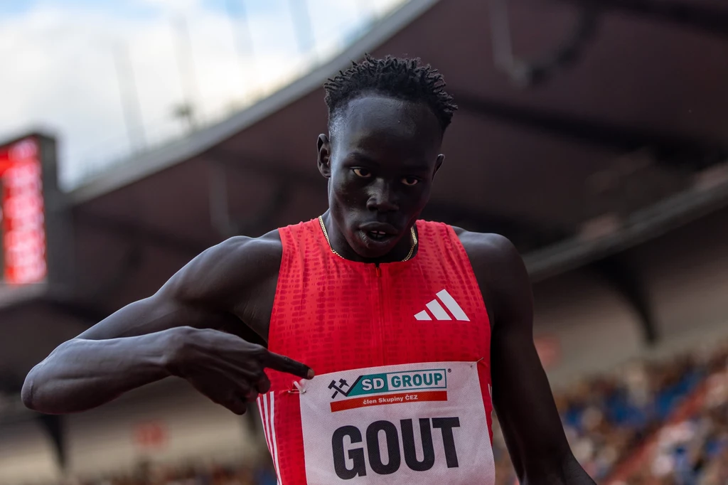 Gout Gout Lekkoatleta w czerwonym stroju sportowym wskazuje na swój numer startowy podczas zawodów na stadionie, w tle widownia i elementy stadionu.